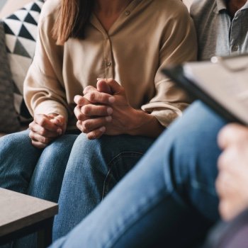 Couple seen sitting, holding hands in love and comfort on a couch with their therapist across from them on a chair with a pen and pad.